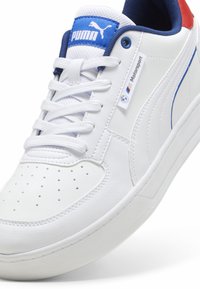 Sneaker in pelle bianca con accenti blu e rossi, punta perforata, lacci bianchi e una patch con logo che presenta il marchio e dettagli motorsport.
