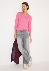 Roze V-hals trui, grijze wijde jeans, met een kastanjebruine jas in de hand. Beige en blauwe sneakers maken de look af met een casual stijl.