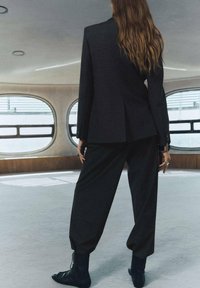Czarny dopasowany garnitur z obcisłą marynarką i luźnymi spodniami, w połączeniu z czarnymi butami na sznurówki; gładki materiał i elegancka sylwetka.