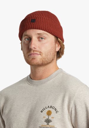 Gorro de punto de color óxido con textura acanalada, que presenta un parche de logo negro. Llevado con una sudadera gris claro que muestra detalles impresos.