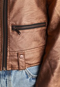 Veste en cuir bronze avec poche zippée, surface texturée et fermeture à pression. Associée à un jean en denim bleu clair usé.
