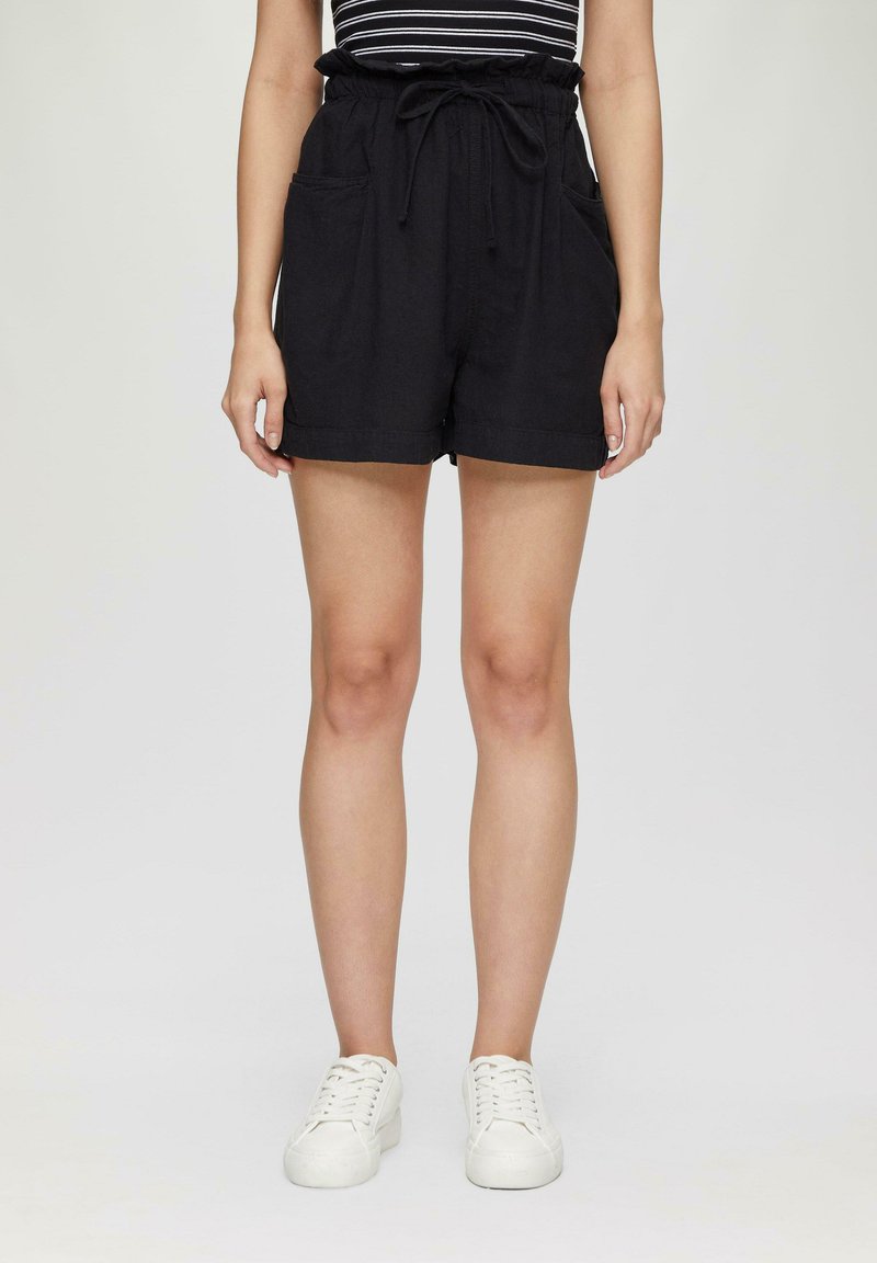Shorts en lin noirs avec un waist à cordon, deux poches avant et une coupe décontractée. Associés à des baskets blanches, soulignant un look décontracté.