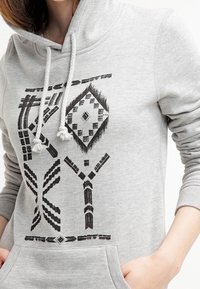 Sudadera con capucha gris claro con patrones geométricos negros en la parte delantera. Cuenta con capucha ajustable con cordón, bolsillo lateral tipo canguro y puños acanalados.