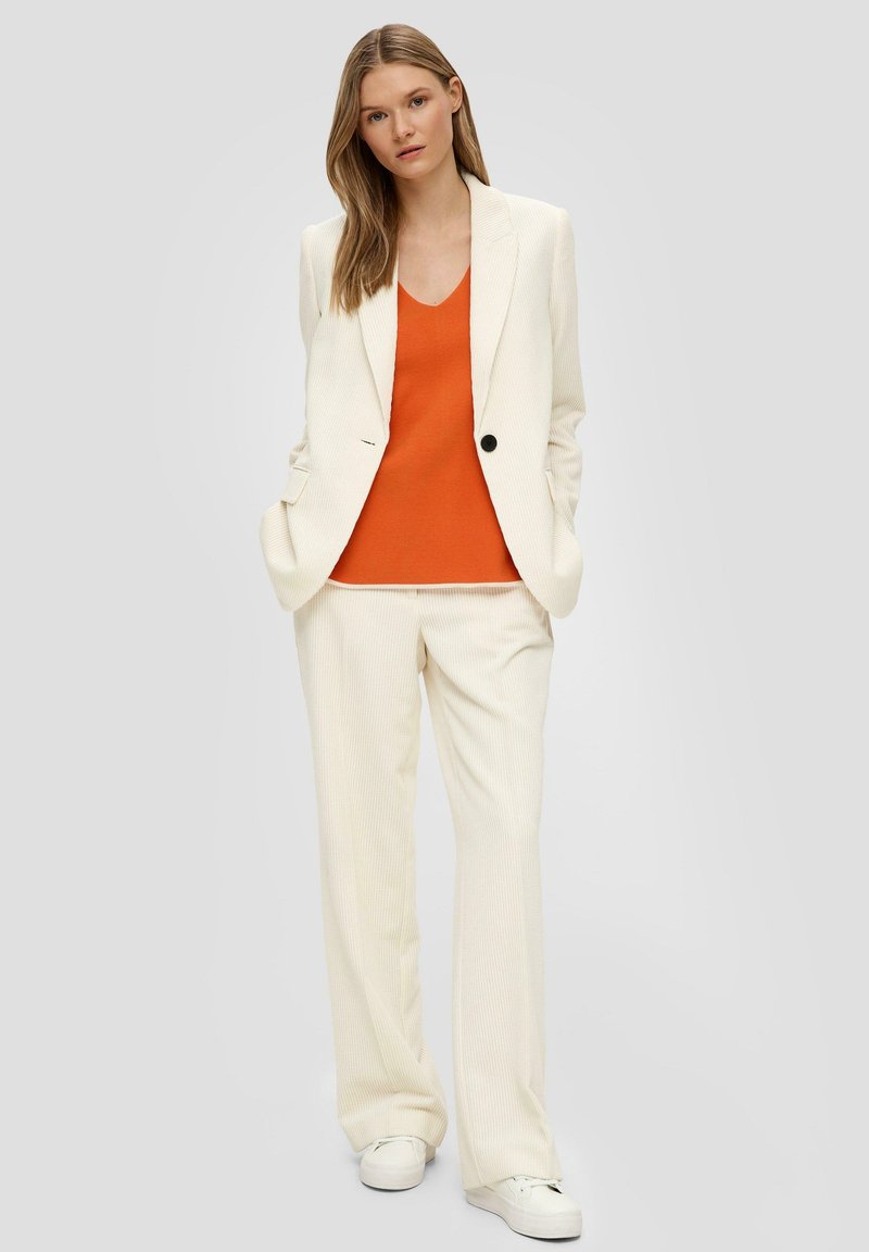 Witte, geribbelde blazer met één zwarte knoop, gecombineerd met een gestructureerde oranje top en bijpassende witte broek met wijde pijpen.