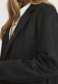 Blazer in misto lana nero con rever a punta e maniche lunghe. Il tessuto ha una texture morbida, con una vestibilità comoda e dettagli minimi.