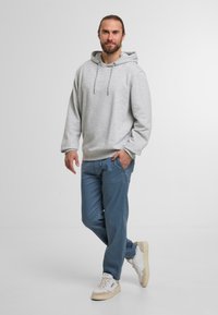 Grauer Kapuzenpullover mit Kordelzug, kombiniert mit blauen Sweatpants und cremefarbenen Sneakers. Das Model steht lässig mit einer Hand in der Tasche.