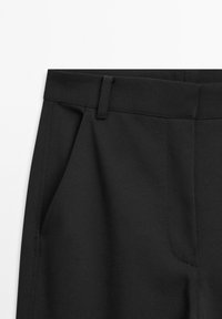Černé tailored kalhoty s hladkým povrchem, mají plochý přední díl, poutka na opasek a boční kapsy. Švy jsou elegantní a minimalistické.