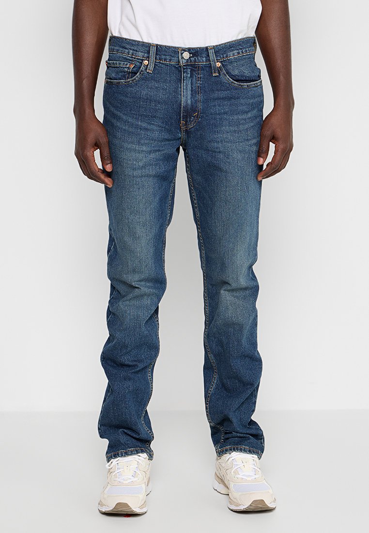 Personne portant un jean droit bleu et des baskets blanches se tenant devant un fond blanc uni.