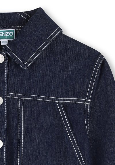 Veste en denim bleu foncé avec un col pointu, des coutures blanches et des boutons-pression argentés. Comprend une poche poitrine et des fentes latérales.