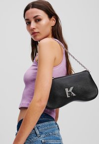 Borsa a mano in pelle nera con forma curvata, dotata di una tracolla in catena argentata e un dettaglio con logo. Il modello indossa un top lavanda e pantaloni shorts in denim.