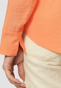 Chemise longue à manches longues orange froissée avec une coupe décontractée, présentant des plis au niveau des poignets et un ourlet arrondi, associée à un pantalon couleur crème.