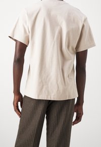 Henkilö, jolla on lyhythihainen beige t-paita ja ruskeat raidalliset housut, näkyy takaa tasaisella valkoisella taustalla.