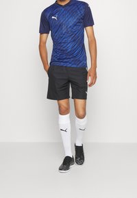 Puma sportkläder med en mörkblå mönstrad kortärmad tröja, svarta shorts, vita knästrumpor och svarta fotbollsskor.