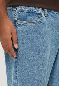 Ανοιχτό μπλε τζιν με κλασική εφαρμογή, με χάλκινα καρφιά, μπροστινή τσέπη και διακριτή υφή από το τζιν ύφασμα.