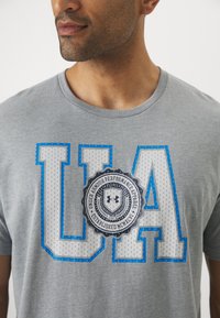 Grå bomulds t-shirt med stort "UA" print i blå og grå, med et cirkulært logo-design nedenfor, rund hals og korte ærmer.