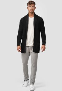 Cárdigan negro de punto con cuello chal, textura acanalada y bolsillos frontales, combinado con pantalones gris claro y zapatillas blancas.