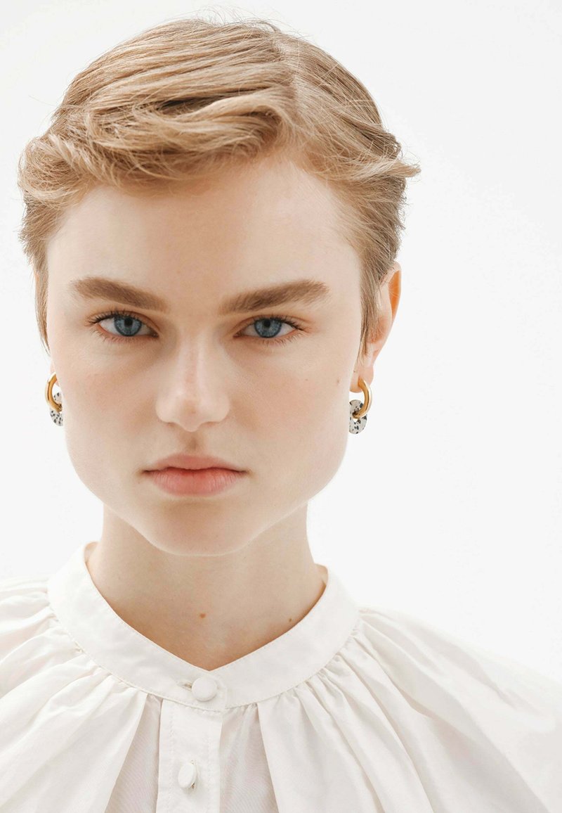 Camisa blanca de botones con cuello plisado; cabello rubio corto y peinado; grandes pendientes de oro y negro; ojos azules claros.