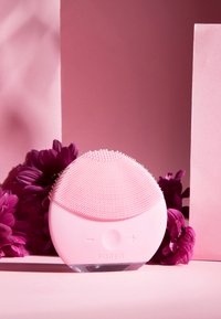 Siliconansiktsrengöringsapparat i ljusrosa, med texturerade borst på toppen, kontrollknappar och en genomskinlig bas. Omgiven av lila blommor.