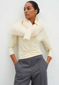 Chunky crèmekleurige sjaal met ribbelstructuur, losjes om de nek gedrapeerd, gecombineerd met een aansluitend lichtgele top en grijze broek.