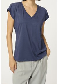 Donna che indossa una maglietta blu navy a maniche corte con scollatura a V, leggermente infilata in un pantalone grigio chiaro a vita alta, con una mano in tasca.