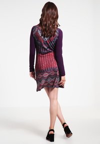 Multikleurige jurk met een geometrisch en stippenpatroon. Lange paarse mouwen, een getailleerde lijfje en een uitlopende rok. Zwarte hoge hakken.