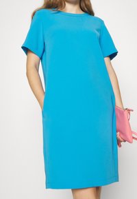Modré šaty krátkého rukávu z hladké látky, s kulatým výstřihem a bočními kapsami. V jedné ruce drží malou růžovou tašku.
