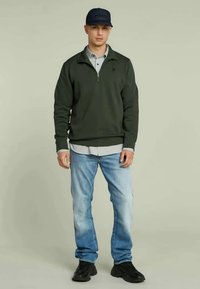 Felpa zipata verde scuro, indossata sopra una maglietta grigia, abbinata a jeans azzurri chiari e sneakers nere. Il modello è in piedi contro uno sfondo neutro.