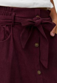 Jupe en velours côtelé bordeaux avec une ceinture nouée à la taille, dotée d'une fermeture à boutons et de deux poches latérales. Le tissu a une texture côtelée.