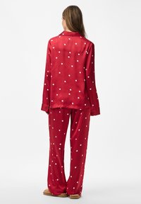 Femme debout portant un pyjama rouge à manches longues avec des motifs de coeurs blancs et des chaussons beiges, vue de dos sur fond blanc.