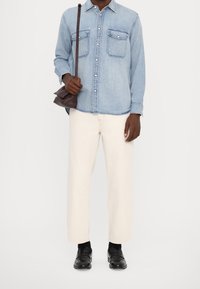 Camisa de mezclilla azul claro con dos bolsillos en el pecho, pantalones holgados de color blanco roto y mocasines negros, complementados con un bolso marrón cruzado.