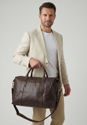 Mand iført beige blazer og hvid skjorte, der holder en stor brun læder rejsetaske med top håndtag og skulderrem.