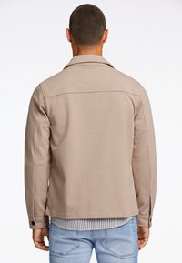 Giacca beige chiaro realizzata in tessuto texturizzato, con un colletto su misura e polsini con bottoni, abbinata a una camicia a righe sottostante.