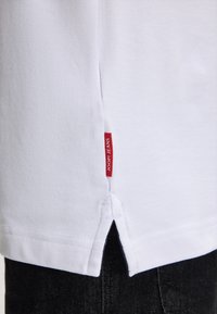 Witte katoenen T-shirt met een zijscheurtje aan de onderrand en een rode "JOOP! JEANS" label dicht bij de onderkant, gedragen met donkere jeans.