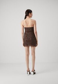 Robe mini marron avec fines bretelles, ornée de motifs en spirale blancs complexes et de sequins. Le modèle porte des sandales à talons carrés. Vue de dos présentée.