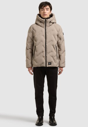 Jeune homme debout portant une veste matelassée à capuche beige, un pantalon noir et des bottes noires sur un fond clair uni.