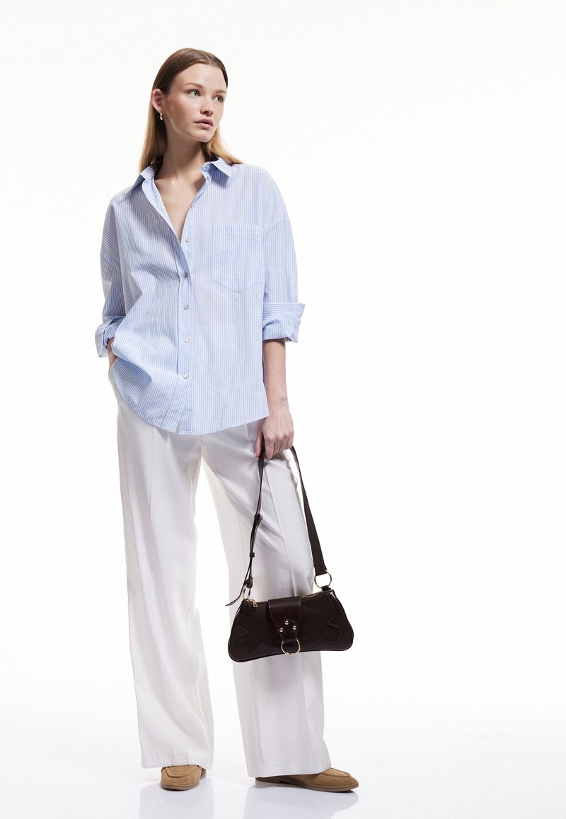 Camisa de botones de rayas azul claro con mangas remangadas, combinada con pantalones de pierna ancha blancos. Bolso marrón con acentos dorados y correa de hombro.