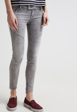 Personne portant un jean skinny gris avec des genoux matelassés et des chaussures slip-on bordeaux, debout devant un fond uni.