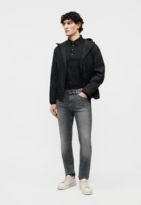 Fiatal férfi fekete kapucnis dzsekiben, fekete galléros pólóban, szürke farmernadrágban és fehér tornacipőben, egyik kezét a zsebében tartva, fehér háttér előtt áll.