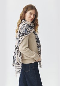 Suéter beige de punto con dobladillo acanalado, acompañado de una gran bufanda en crema y azul marino con un patrón y flecos en los bordes. Jeans de pierna ancha azul oscuro.