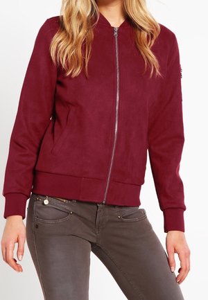 Giacca bomber bordeaux in suede con chiusura a zip, polsini e orlo a coste, e tasche laterali. Design semplice con linee pulite.