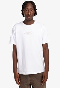 Camiseta blanca de algodón con mangas cortas, que presenta un logo impreso en beige claro "ELEMENT SKATEBOARD CO. WORLD WIDE" en la parte delantera.