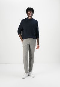 Navy textured knit sweater, light gray trousers, and white sneakers. The model stands against a plain white background.