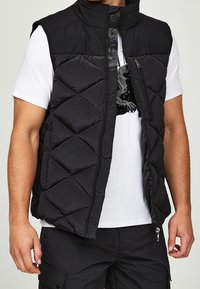 Černá prošívaná vesta s diamantovým vzorem prošití, vysokým límcem a zipem vpředu. Má logo a je nošena přes bílé tričko.