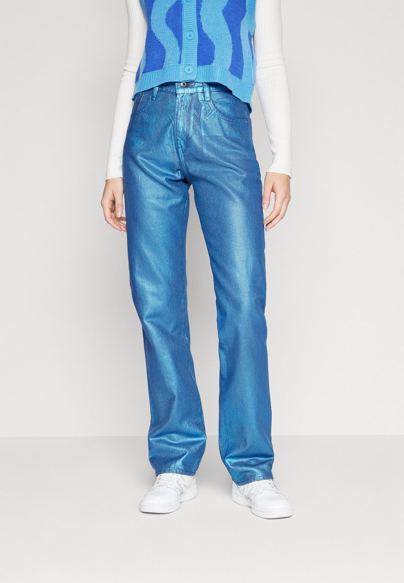 Personne portant un jean droit bleu brillant, des baskets blanches, un haut blanc à manches longues et un pull sans manches à motifs bleus.