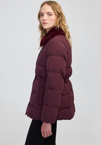 Giovane donna con capelli biondi mossi, che indossa una giacca imbottita bordeaux con colletto di pelliccia, in piedi di profilo su uno sfondo neutro.