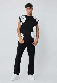 Pull sans manches noir en tricot avec un grand motif floral blanc, associé à un pantalon noir et des baskets blanches. Mains visibles, modèle debout.