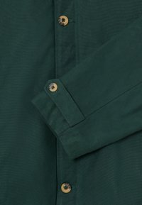 Σκούρο πράσινο μανίκι που είναι διπλωμένο πάνω από το κουμπωμένο μπροστινό μέρος, διακοσμημένο με καφέ και μαύρους μαρμαρωμένους κουμπιά σε βαμβακερό ύφασμα.