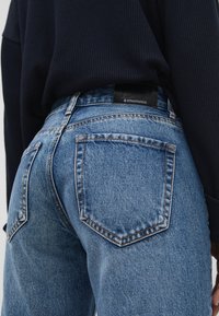 Lähikuva henkilöstä, joka on pukeutunut sinisiin farkkuihin, joissa on takataskut ja musta vyötärötunniste, yhdistettynä tumman väriseen ribattu puseroon.