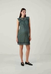 Akyliškas nėrinių suknelė melsvos spalvos, trumpomis nelygiomis rankovėmis ir prigludusia liemene, su gėlių raštu ir banguota apdaila. Dėvima su juodais butais.