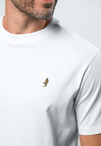 Homme barbu portant un t-shirt blanc uni avec une petite figure brodée représentant une personne en manteau près de la poitrine.